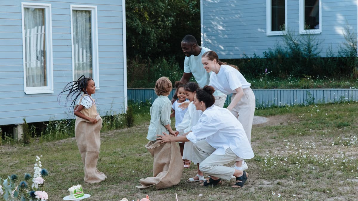 Joyful family gathering with children playing sack race in a garden outside a house.